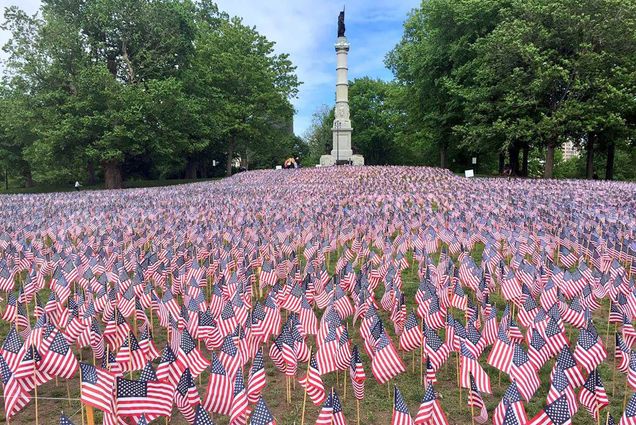 Photo of small American flags planted in the ground around Boston Common, a tall, column like statue is seen in the background as well as trees.