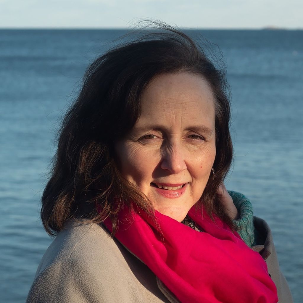 Headshot of Regina Hansen. She wears a jacket and bright, reddish pink scarf, and smiles as the wind blows her hair to her right. The ocean is seen behind her.