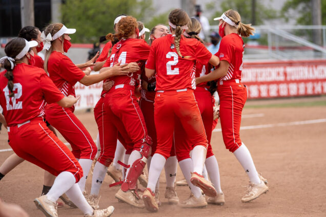 Photo of the BU Softball team jumping in celebration after they won their third straight Patriot League title on Saturday May 15, 2021. The women wear read jerseys, white visors and red face masks.