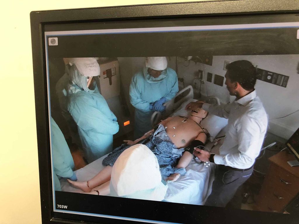 A video monitor displaying a training exercise of the Boston Medical Center Special Pathogens Unit (SPU). A doctor and SPU members in hazmat suits stand over a mannequin dummy patient in a hospital room.