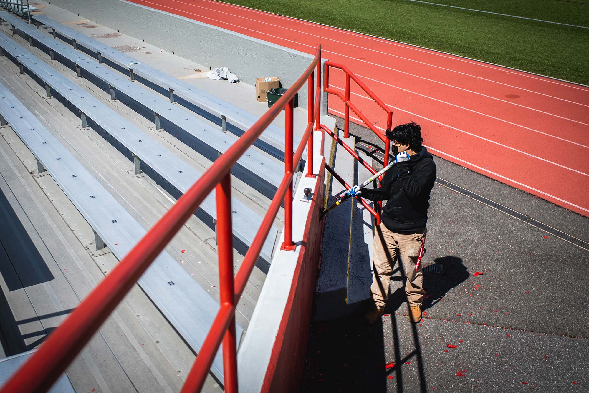 Photo of Kevin Beoya of Wilder Painting in a black sweatshirt, mask, and khaki paints, holding a long stick, and painting a red wall around the bleachers at Nickerson field.