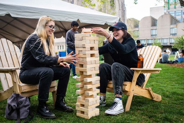 Photo of Eden McColl (SHA 21) and Morgan Michelson (SHA 21) playing a game of Jenga during Senior Week. The pair sit on wooden adirondack chairs, as McColl holds the tower from tipping over and Michelson attempts to place a piece on top. Both students wear black, Michelson wears a Red Sox hat. Behind them, a white tent and other students are seen on the BU beach.