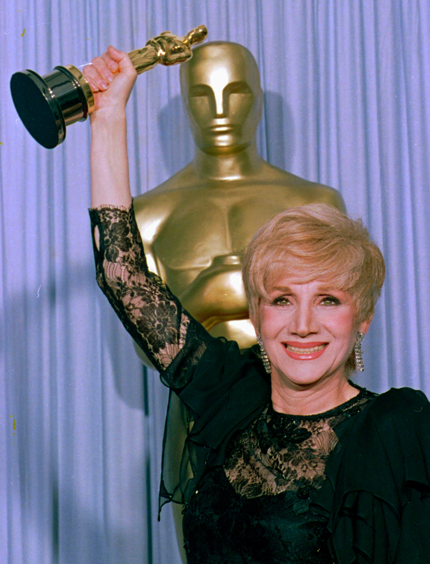 Photo of Olympia Dukakis holding her Oscar at the Shrine Auditorium in Los Angles after being honored at the 60th Academy Awards as best supporting actress for her role in "Moonstrck." She wears a black, lacy gown and smiles brightly. Behind her, a large oscar statue and purple curtain are seen.