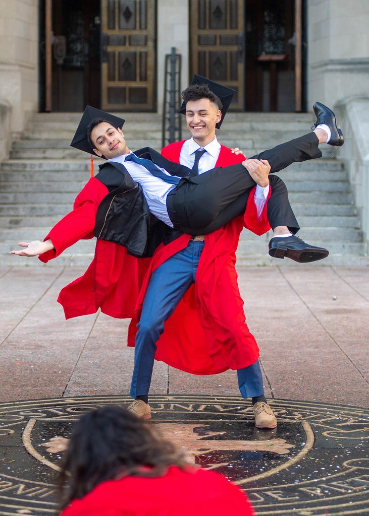 Photo of Mrinal Ghosh (ENG’21) with his arms and legs extended as Camden Kronhaus (ENG’21) holds him up over the seal in Marsh Plaza May 11. They both wear suits and their caps and gowns and smile.