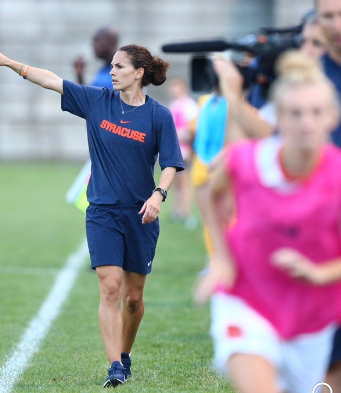 Photo of Kelly in a blue Syracuse shirt and shorts pointing to her right as athletes in bright colors run to her left, the athletes are blurred.