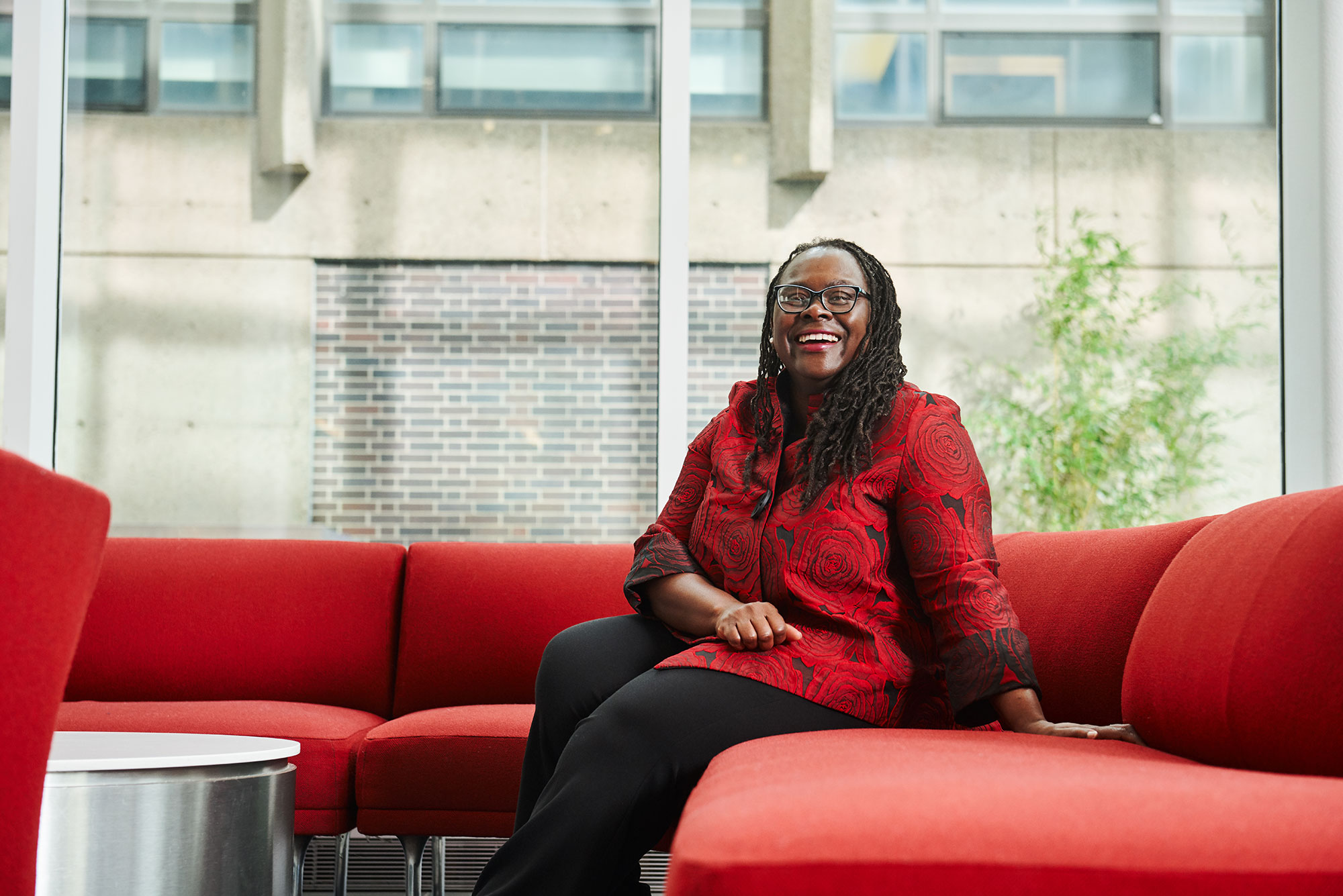 Photo of Law Dean Angela Onwuachi-Willig with a red and black floral blouse sitting on a red couch in and smiling.