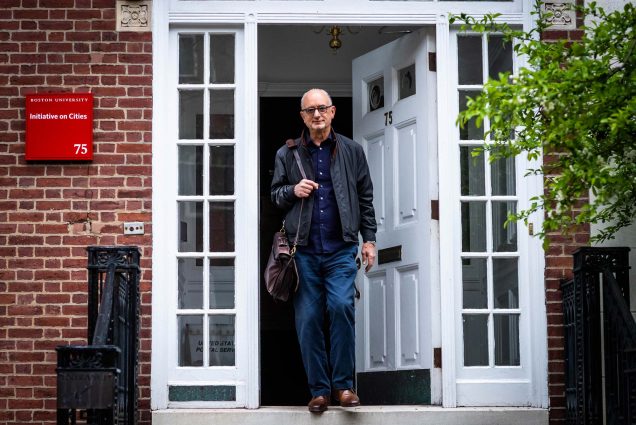 Portrait of Graham Wilson, Founding Codirector of Boston University’s Initiative on Cities, as he walks out of a brick building with a white door and trim with a bag over his shoulder. He wears a black bomber jacket and blue pants. On the left, a red plaque on the building reads “Boston University, Initiative on Cities, 75”