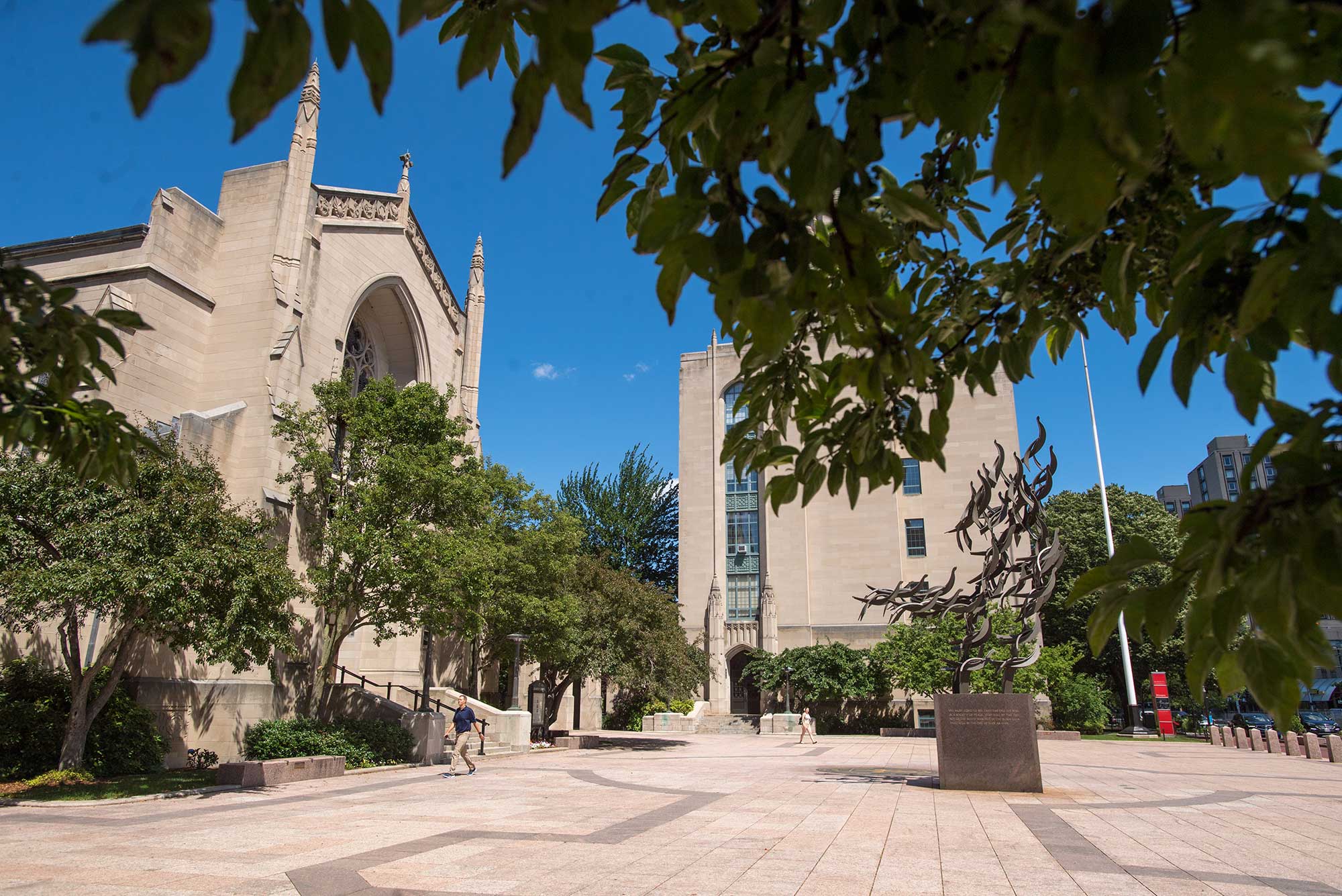 Photo of Marsh Plaza from the west side looking towards the MLK statue with the chapel at left. The sky is blue and the trees are filled with green leaves.