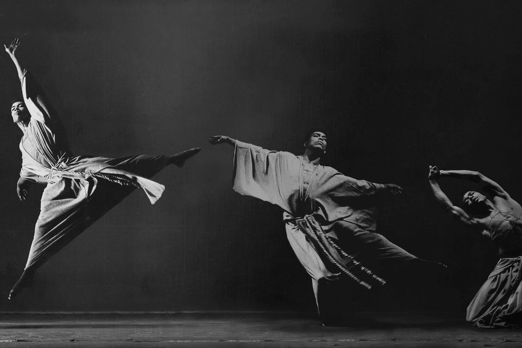Black and white photo of a Black dancer in three different poses, on the left he jumps and stretches his arms and legs, in the middle he extends his arms, on the right he kneels and extends his arms over his head.
