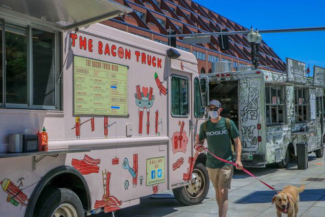 Photo of the “The Bacon Truck,” a pink food truck decorated with cartoon bacon strips. A man with a green shirt, baseball hat and face mask walks by with his dog.