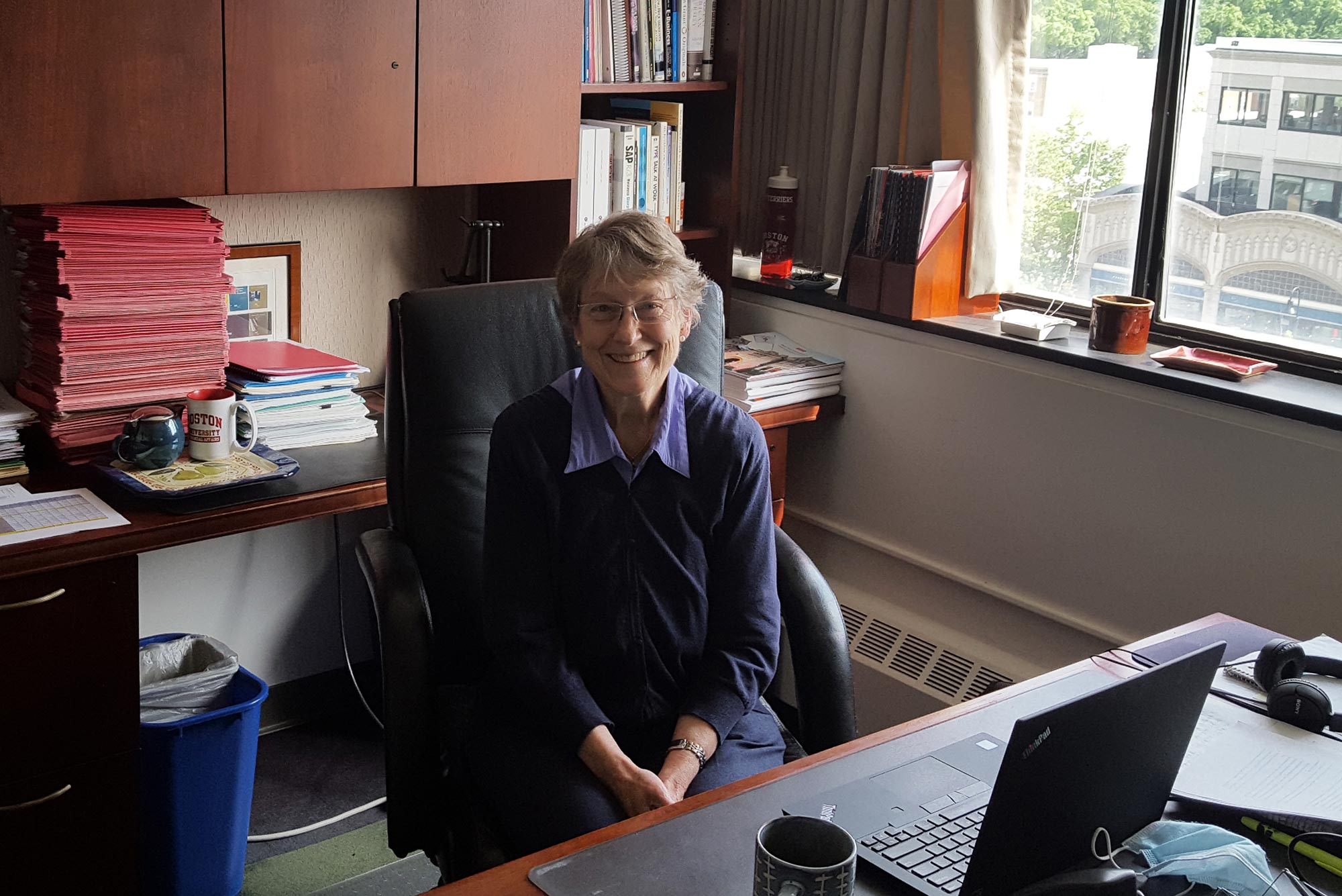 Photo of retiring BU comptroller Gillian Emmons sitting at her desk. She wears a blue sweater, and smiles; a window behind her looks out into Boston. A stack of red booklets sits on the desk behind her.