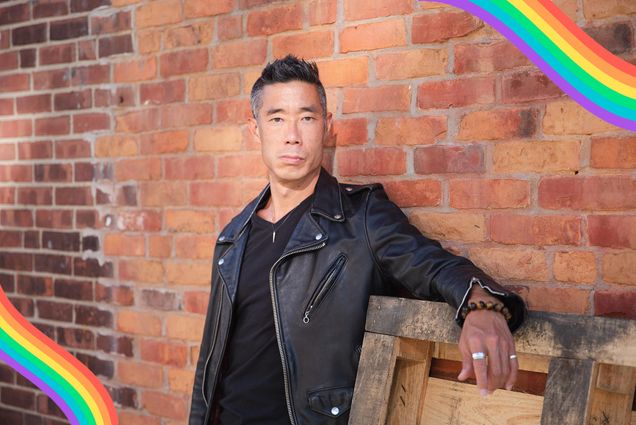 Photo of Kính with a black leather jacket and black shirt leaning against a wooden pallet that is balanced against a brick wall. Pastel rainbow overlays are overlaid on the bottom left and top right.