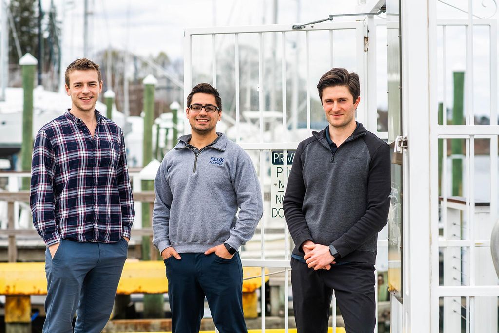 Photo of Flux Marine founders Jonathan Lord (from left), in a blue and white plaid shirt with his hands in his pockets, Ben Sorkin, in a gray quarter zip and glasses and his hands in his pockets, and Daylin Franton, in a dark gray quarter zip holding his hands in front of him, standing on a dock in East Greenwich, R.I. In the background, blurred docked boats can be seen.