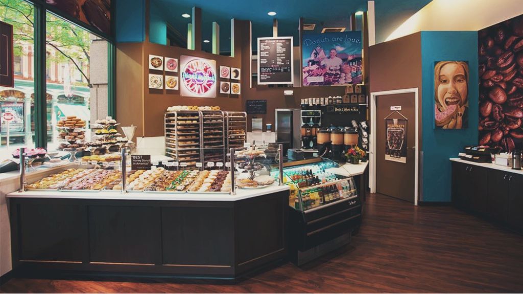 Photo: Kane's Donut's in Boston, MA, a well-lit donut shop interior showcasing a vibrant selection of donuts in a glass display case. To the right, a beverage station is equipped with an array of syrup bottles and a coffee machine. The shop’s menu board is prominently displayed above the counter, and additional baked goods are arranged on racks to the left. Large windows fill the space with natural light, complemented by donut-themed art and warm, chocolate-toned wall panels.