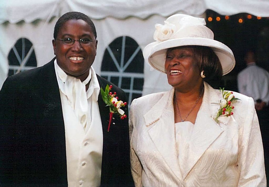 Burgess (left) with her mother on Burgess’ wedding day in 2004. Burgess wears a black tuxedo and her mom wears a white blazer and fancy hat; both smile wide.