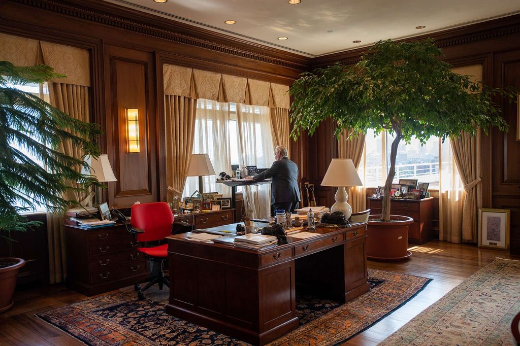 Photo of President Robert A. Brown taken from slight behind, from the other side of his office. He looks at at a laptop on standing desk, and wears a blue suit. His office is large and wood-paneled, has a stately wooden desk, with two large tree-like plants on either side. Long gold curtains hang at the sides of the windows letting sunlight in.
