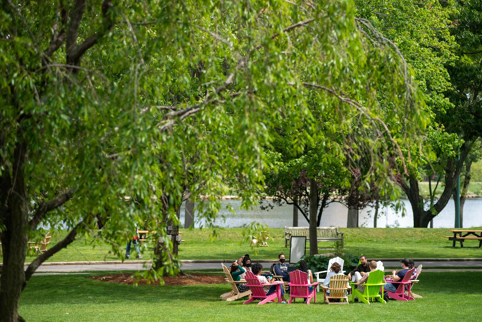 Photo of a group of students, some masked, other unmasked, sitting outside on BU beach in colorful Adirondack chairs. In the foreground, a bright green tree is seen and the day looks sunny and bright.