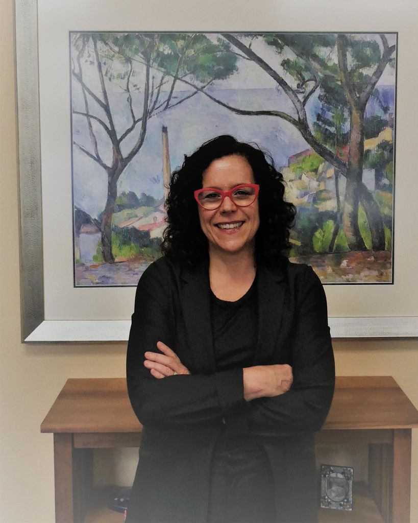 photo of Lynn O’Brien Hallstein in a black blazer and shirt, wearing red glasses, with her arms crossed as she smiles. Behind her, is a pastel-colored painting of a countryside.