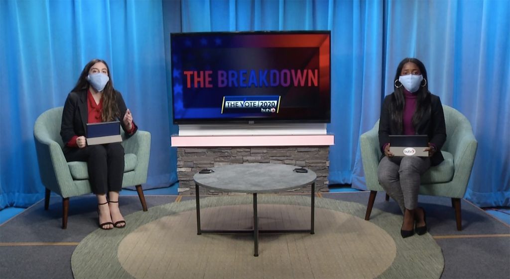 Screenshot from a BUTV10 broadcast, Sophie Falkenheim (COM’22), left, and Archelle Thelemaque (COM’21) were analysts for BUTV10’s The Vote 2020. Both wear light blue face masks and blazers, and sit on two teal chairs on opposite sides of a gray coffee table with a tv behind it that says “The Breakdown: The Vote 2020” on it. Behind them blue studio curtains hang.