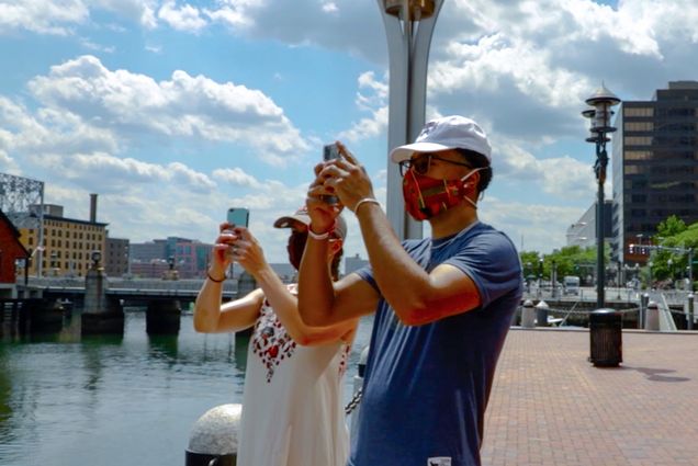 Photo of two people participating in the scavenger hunt; they take a photo of something in the harbor off to their right. Both are wearing face masks and baseball caps.