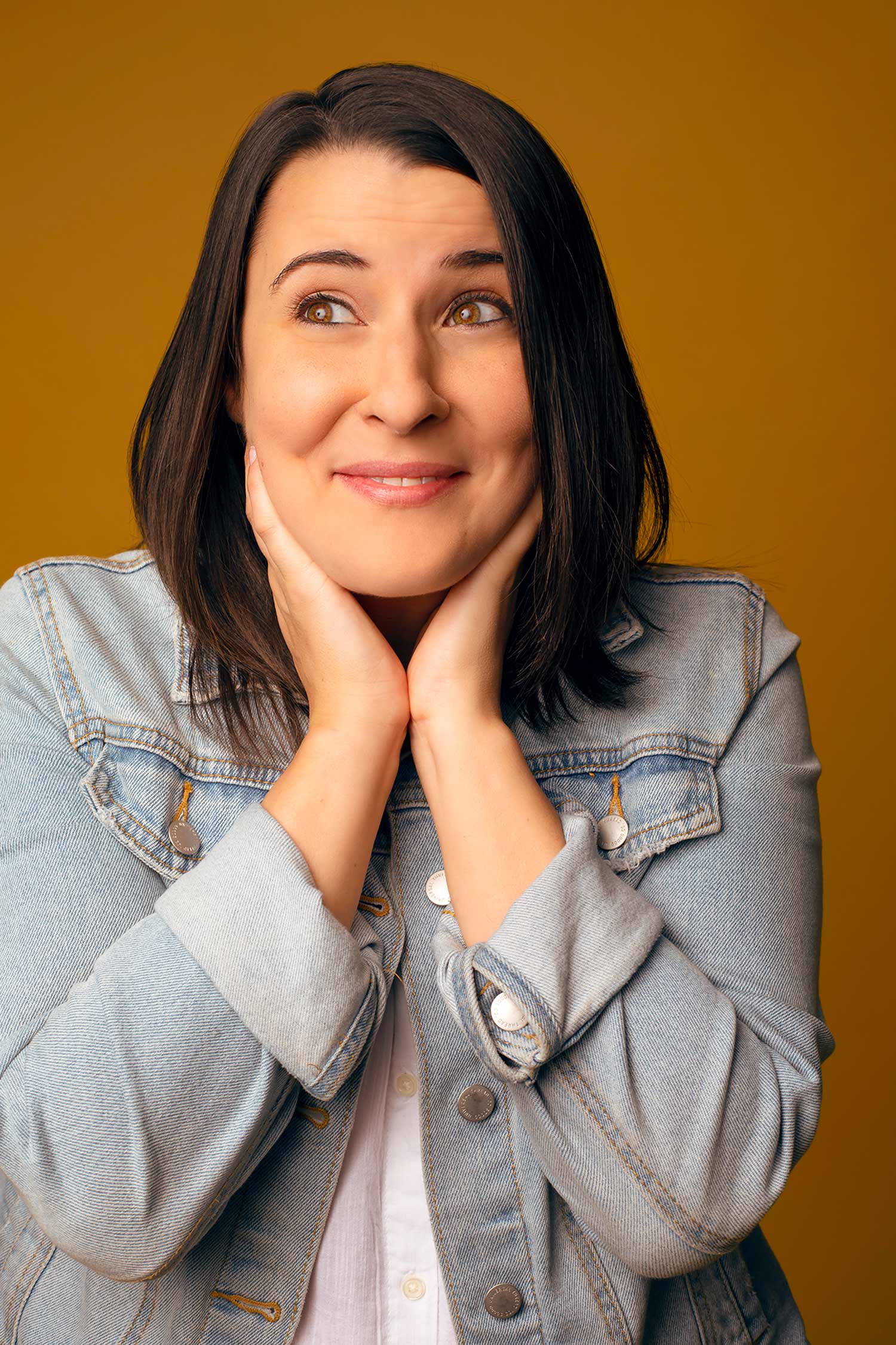 Headshot of Caitlin Durante (COM’14), the cocreator and cohost of the feminist podcast 'The Bechdel Cast', wearing a jean jacket and smiling with her hands on the side of her face. The background is goldish, orange.