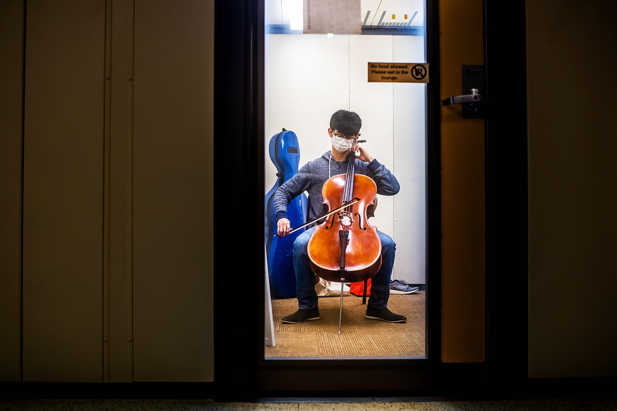 Photo taken through the glass door of a practice room in which Minyung Suh (CFA 22) practices the cello on Monday, July 19, 2021. The outside hallway is dark. Suh wears a white face mask, blue hoodie and jeans.