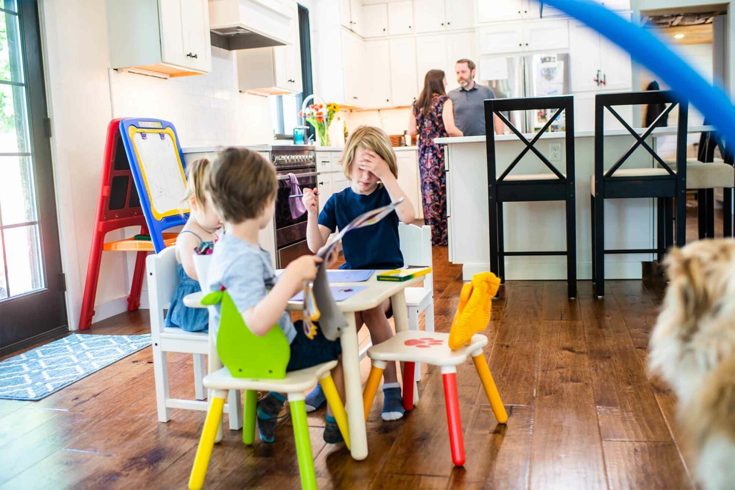 3 children playing or drawing at a kids table while parents talk behind them in the kitchen.