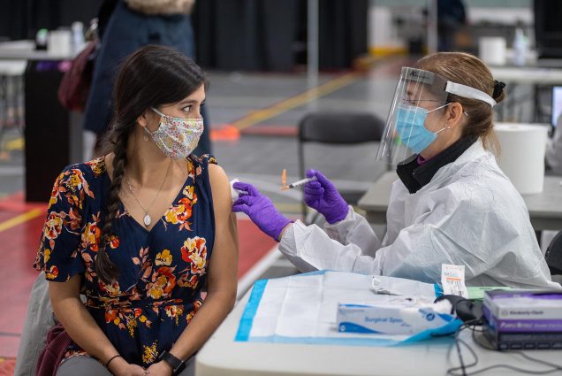 Photo of Anne Herrera (Wheelock’19), triage specialist at Healthway, wearing a floral face mask and blue floral blouse, receiving the Moderna COVID-19 vaccine from Student Health Services Director of Nursing Anne Patey, MSN, at the vaccine clinic January 15 at FitRec. Patey wears gloves, a face mask and face shield.