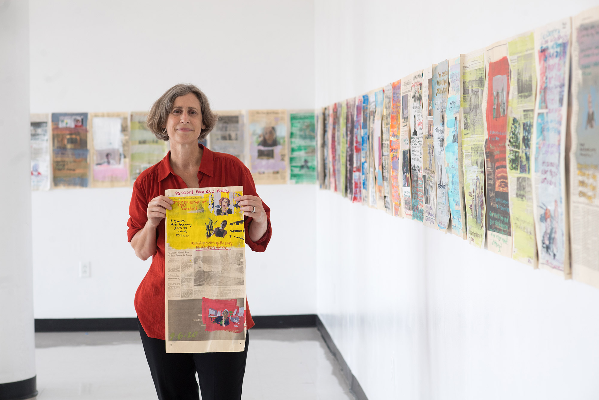 Photo of CFA SVA Director Dana Clancy in a red button down looking towards the camera as she holds a painted page of the NYTimes in a gallery of her work.