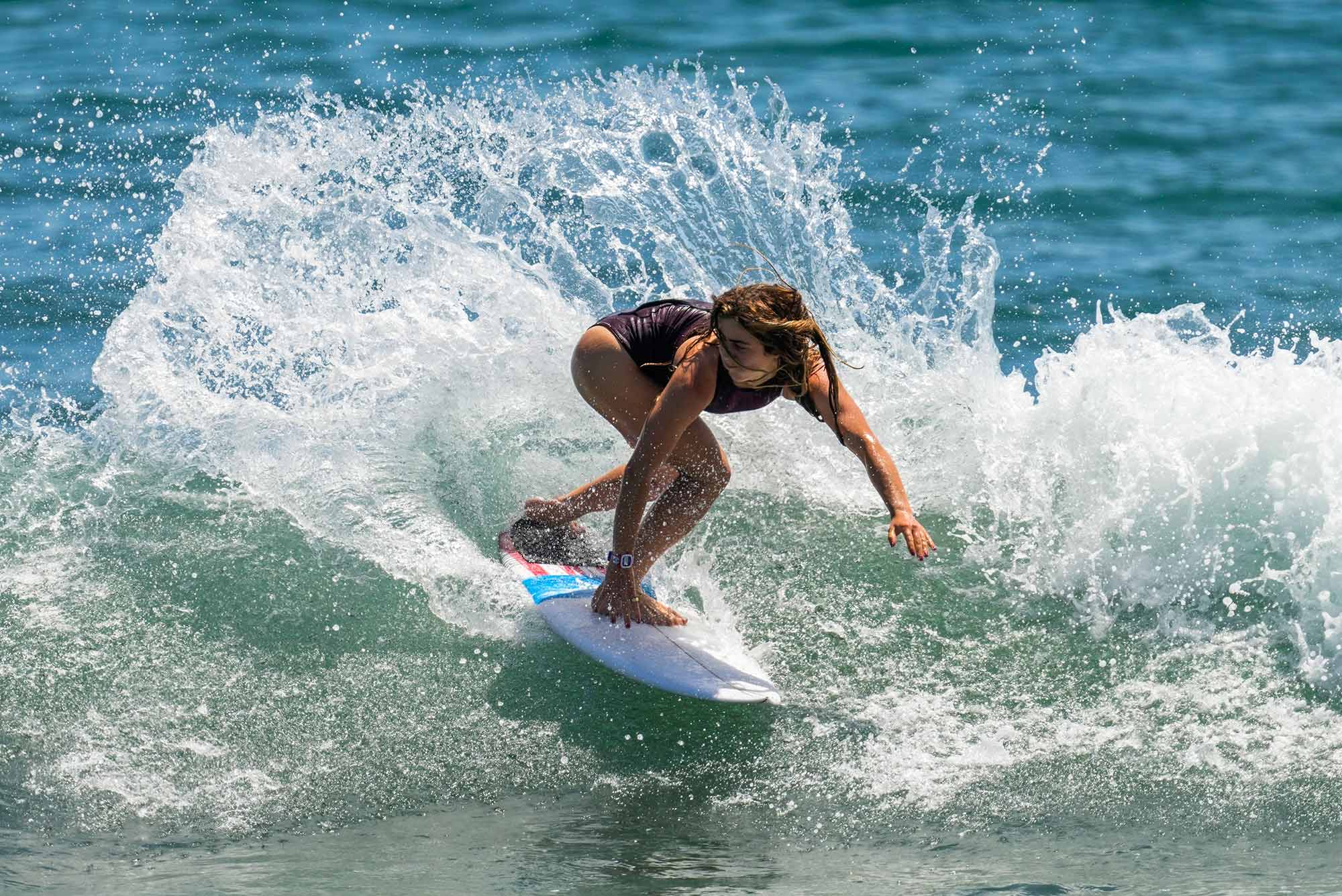 Caroline Marks, of the United States, rides a wave on a white surfboard during a training session at the 2020 Summer Olympics, Thursday, July 22, 2021, at Tsurigasaki beach in Ichinomiya, Japan. The wave splashes up behind her as she crouches on the board.