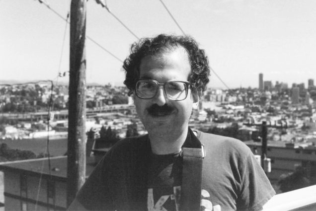 Black and white photo of Laurence A. Bloom (DGE’66, CAS’68). He appears to be on a rooftop and wears some sort of strap over his shoulder.