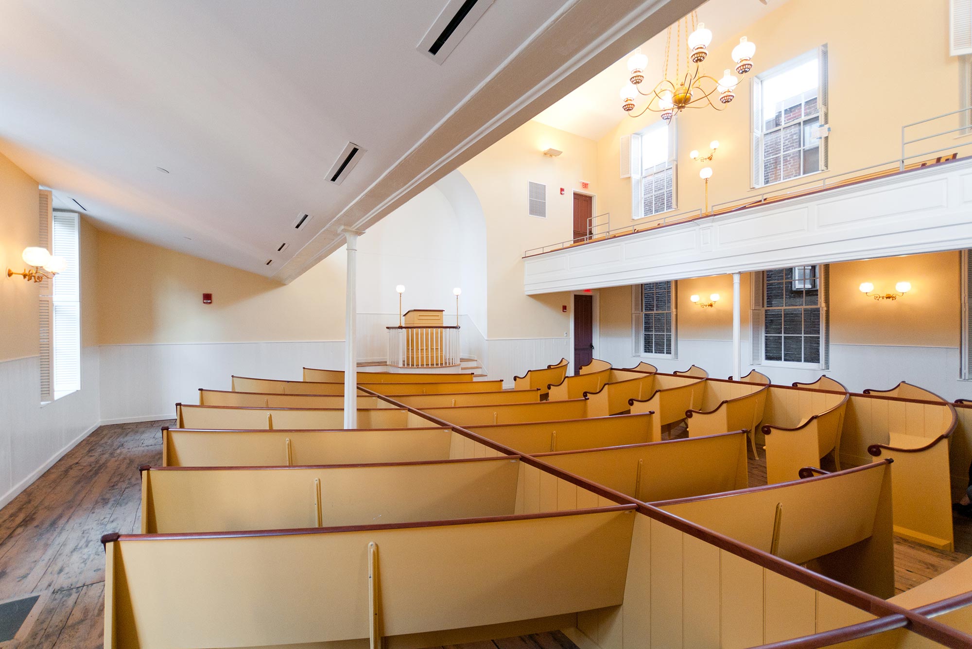 Photo inside the African Meeting House on Beacon Hill, it has wooden pews and a pulpit at the front, with balcony seating on the sides and a chandelier with electric lights.
