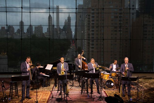 Photo of The Jazz at Lincoln Center Orchestra Septet. A group of 7 men in tuxedos play, the NYC skyline is seen through a window behind them.
