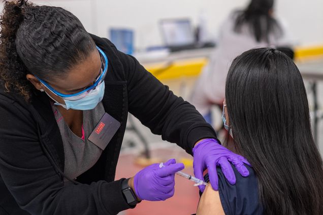 Photo, from behind, of a nurse with blue glasses, wearing a face mask and purple gloves, giving a BU staff or faculty a COVID-19 vaccine, at the vaccine clinic at FitRec January 2020.