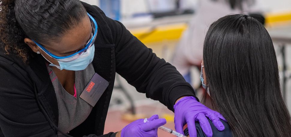 Photo, from behind, of a nurse with blue glasses, wearing a face mask and purple gloves, giving a BU staff or faculty a COVID-19 vaccine, at the vaccine clinic at FitRec January 2020.