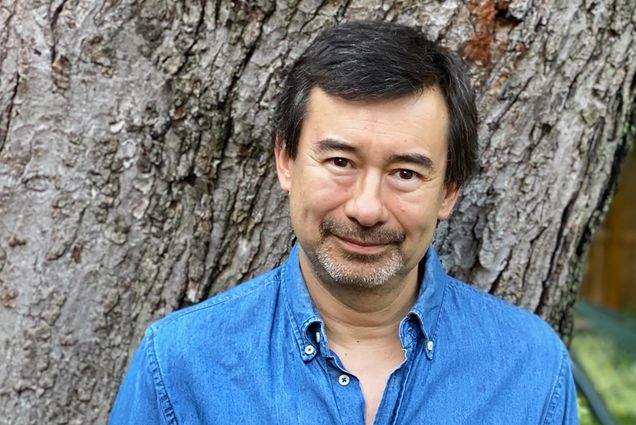 Portrait of Peter Ho Davies in a jean button down shirt in front of a tree trunk.