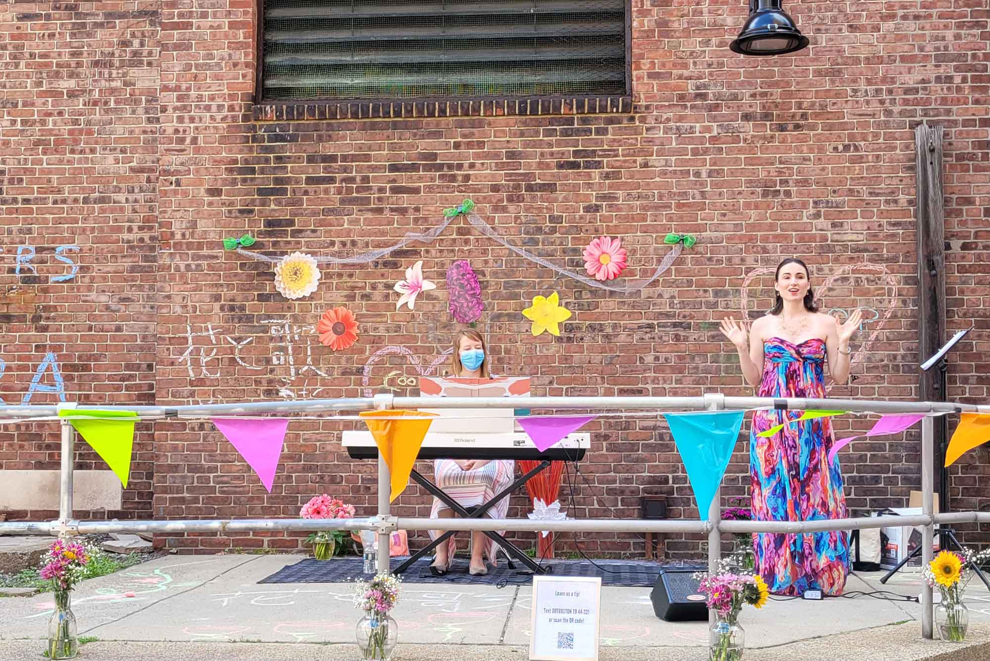Photo of two women, one seated at a white keyboard, the other standing in a colorful dress with her arms extended, perhaps singing. Behind them, a brick wall decorated with flowers is seen.