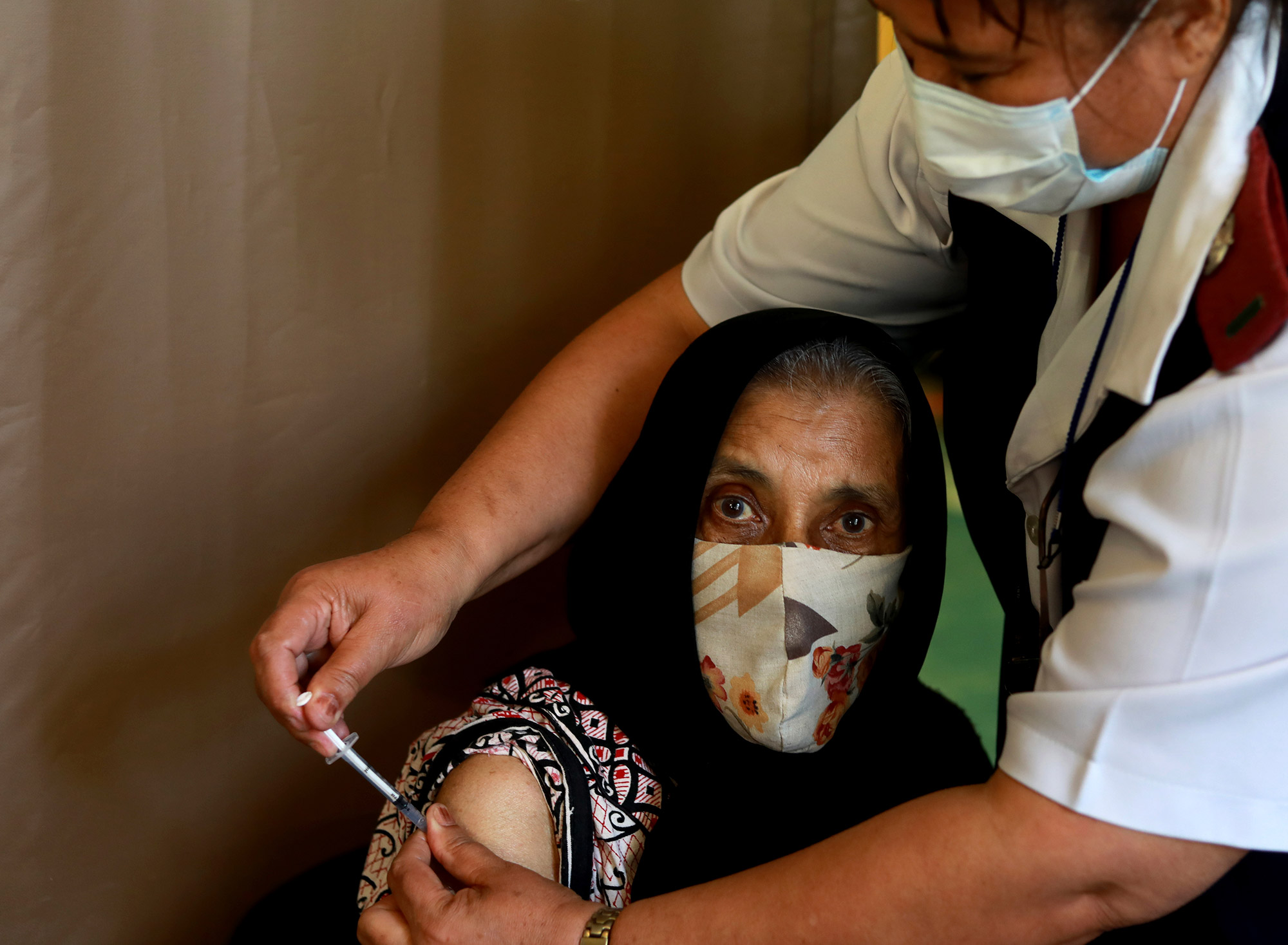 Photo of a seventy six year old woman Kaironesa Sheik Suleman Allie receiving her first dose of the Pfizer COVID-19 vaccine from a health staff member, at a vaccination centre, in Karl Bremer Hospital, in Cape Town, South Africa, Wednesday, May 26, 2021. The staffer wears a blue paper facemask and reaches over her to give the vaccination.