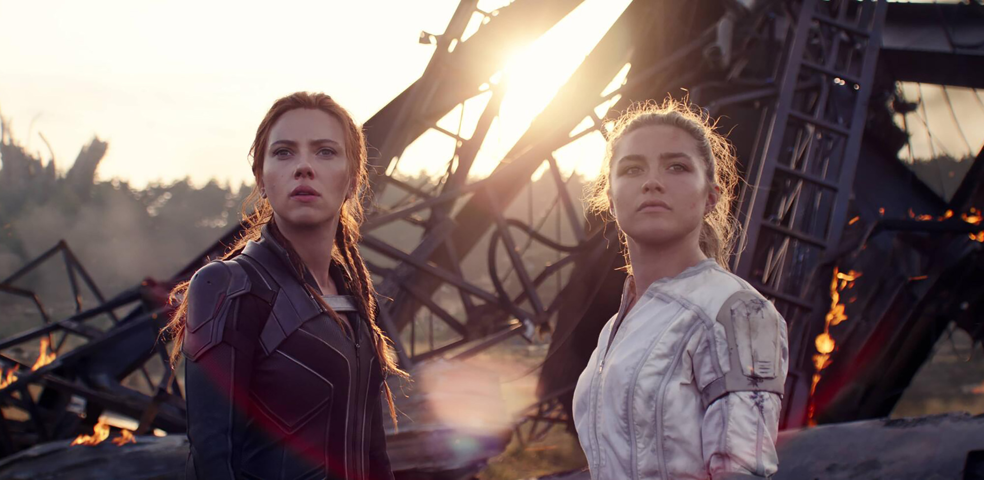Movie still from Black Widow, starring Scarlett Johannson as Natasha Romanoff/Black Widow, at left, wearing a black leather jacket, next to Florence Pugh playing her little sister, Yelena Belova. Behind them, some rubble is seen in the distance.