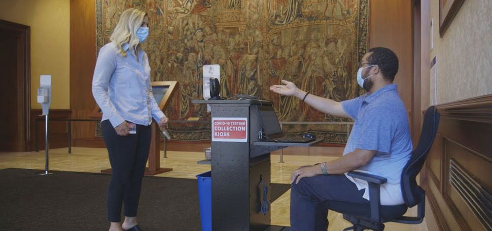 A male staff member at a COVID-19 Test Collection Kiosk prompts a woman to leave her testing swab vial in a tray on the kiosk table.