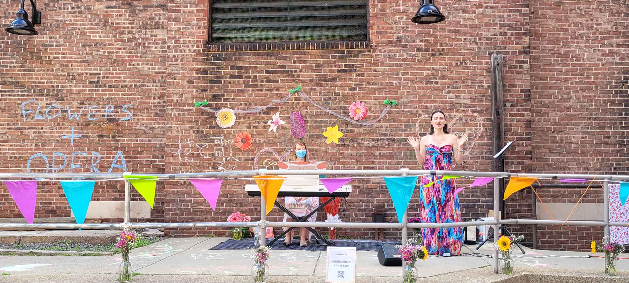Photo of two women, one seated at a white keyboard, the other standing in a colorful dress with her arms extended, perhaps singing. Behind them, a brick wall decorated with flowers is seen.