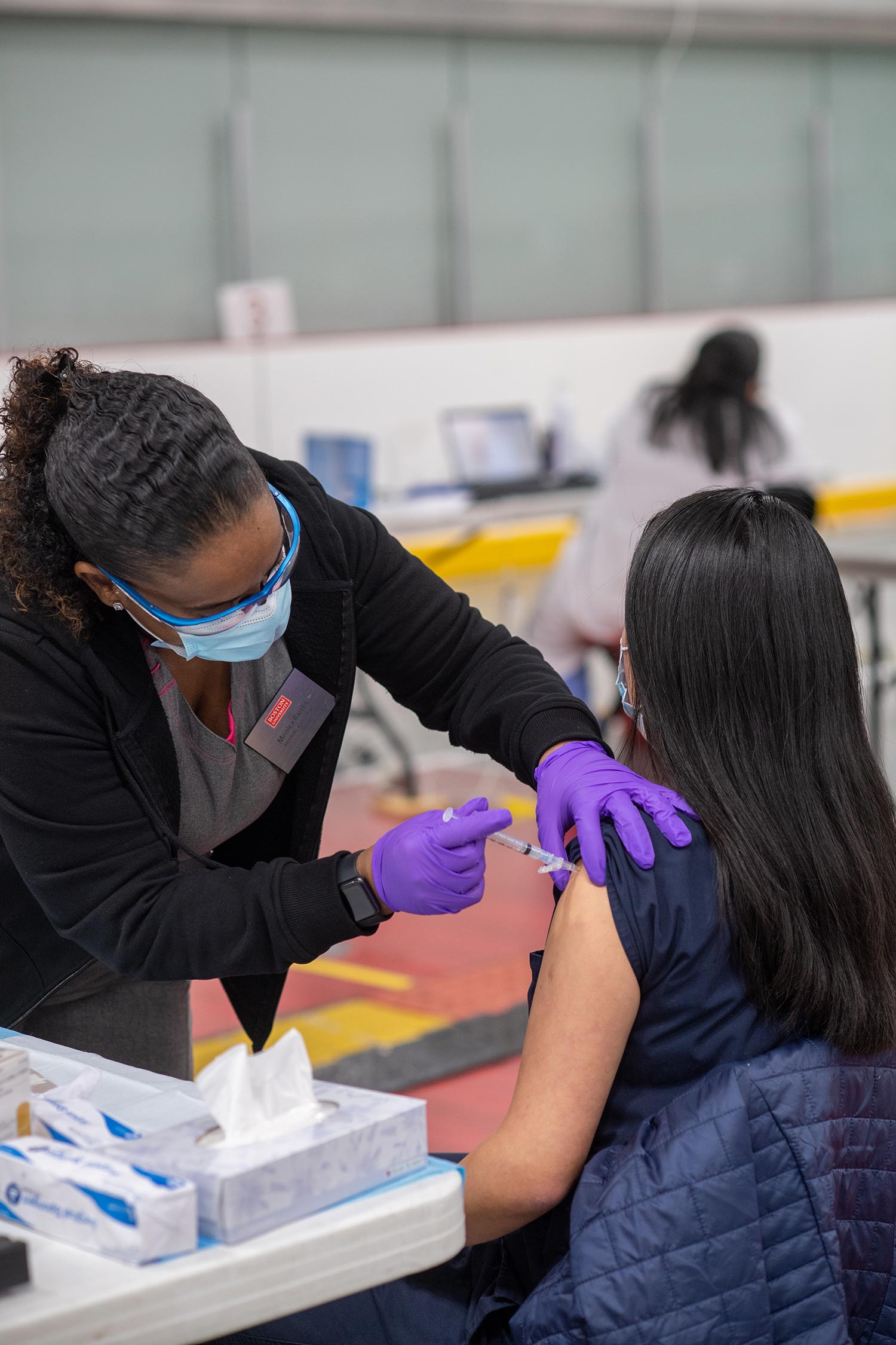 Photo, from behind, of a nurse with blue glasses, wearing a face mask and purple gloves, giving a BU staff or faculty a COVID-19 vaccine, at the vaccine clinic at FitRec January 2020.