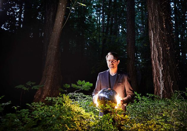 Photo of Aaron Rasmussen (COM’06, CAS’06), with a spherical keyboard he made for himself because of a repetitive strain injury, at home in Washington State. He wears a gray suit jacket, glasses, in stands in the bushes which are illuminated by the lights of his keyboard.