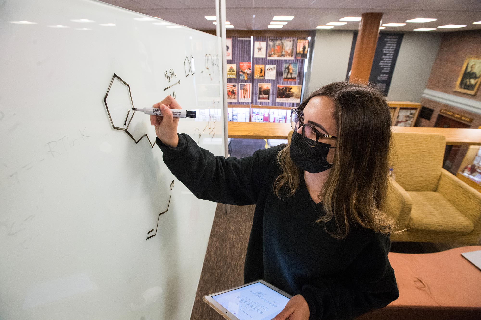 Marian Karam (ENG’22) studies for her Organic Chem1 final June 30, in Mugar Library. She draws chemical bonds on a whiteboard. She wears a black face mask, black sweatshirt and has shoulder length hair.