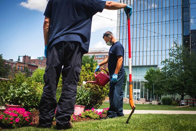 Photo of Chris Tavares of BU Facilities Management and Operations watering some flowers. Another facilities worker in blue and black stands in the foreground with a red shovel in his hand.