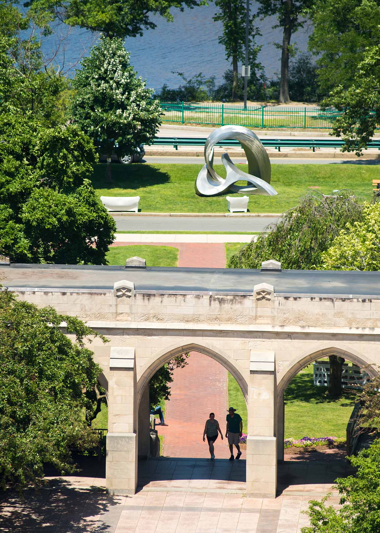 Marsh Plaza and BU Beach viewed form the seventh floor of Photonics. Two people seen in silhouette walk underneath one of the arches, and a shiny, metal sculpture is seen on the BU beach lawn. It is bright, sunny and summery.
