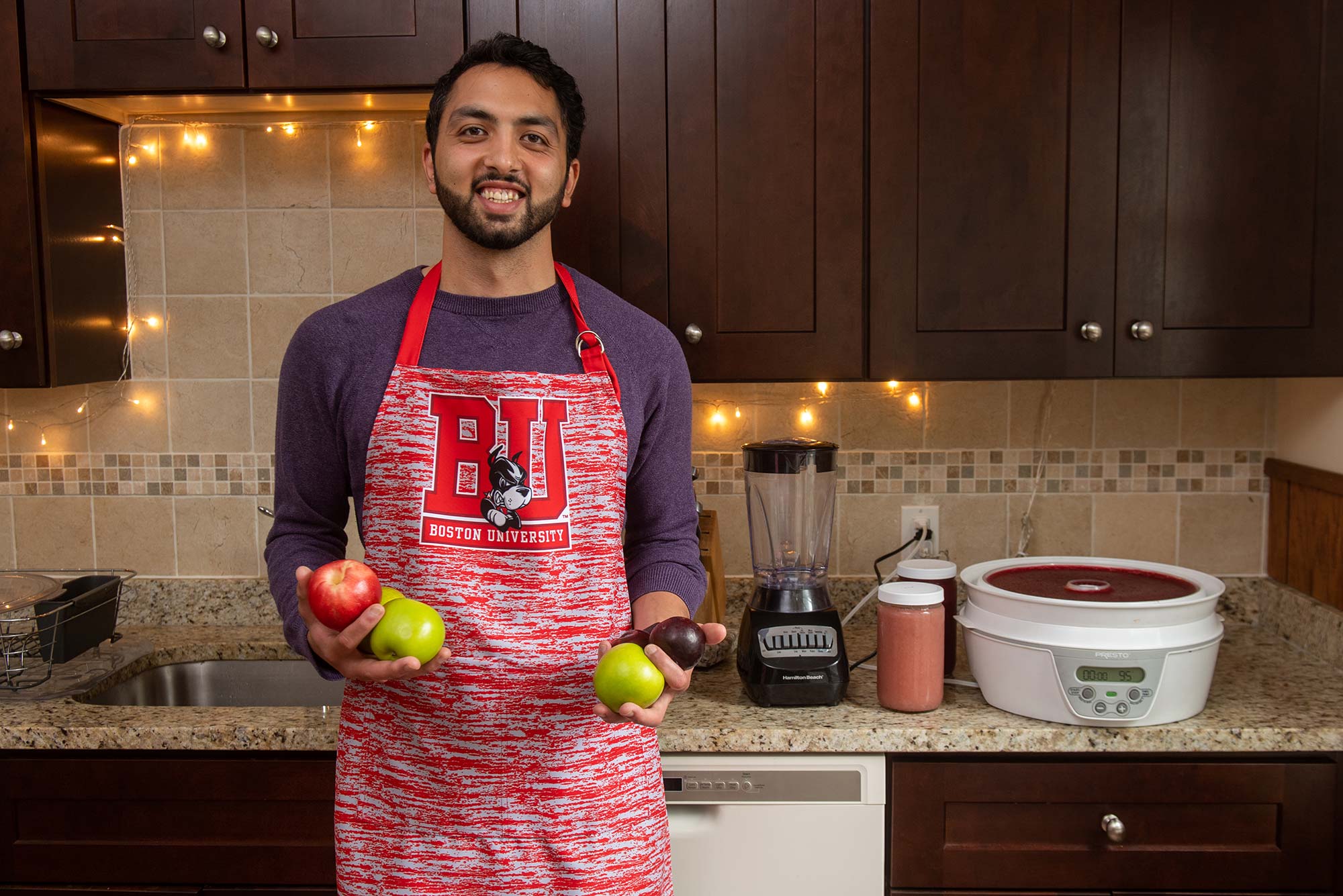 Photo of Andree Entezari, in his Somerville home kitchen, holding various fruits and wearing a white and red-striped BU apron, and smiling. Behind him, a dehydrator and other fruit-leather making tools are seen.