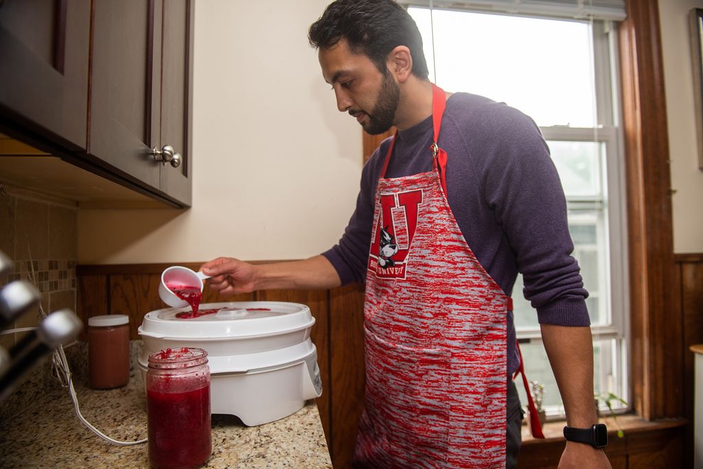 Photo of Andree Entezari, in his Somerville home kitchen, wearing a white and red-striped BU apron, and pouring a reddish fruit concentrate into his dehydrator to make fruit leather.