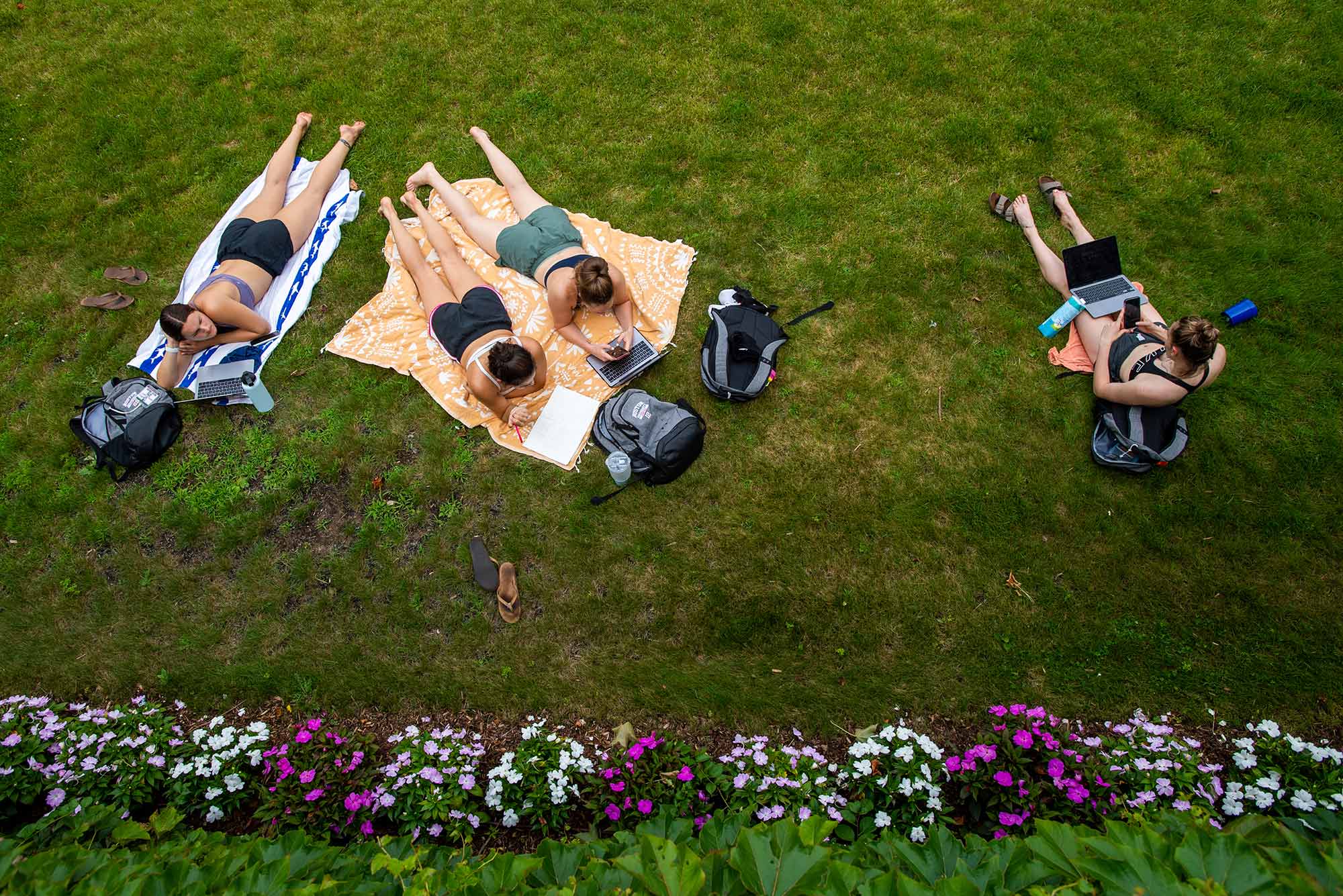 Aerial photo of 7/20/21 members of the BU women’s basketball team, including Riley Childs (COM’22) small forward, from left, Annabelle Larnard (Questrom’23), guard/forward, Emily Esposito (COM’22), guard, and Liz Shean (COM’23), guard, chat on Nickerson Field while working on their summer term class work July 20. The grass is greenish yellow, and there is a row of pink and white flowers in front of them.