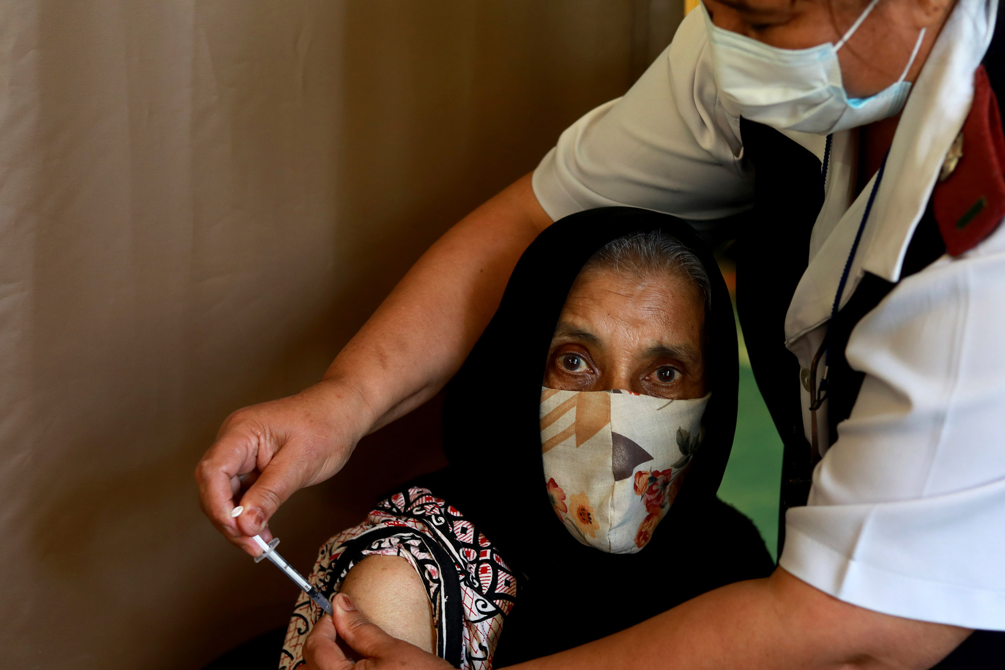 Photo of a seventy six year old woman Kaironesa Sheik Suleman Allie receiving her first dose of the Pfizer COVID-19 vaccine from a health staff member, at a vaccination centre, in Karl Bremer Hospital, in Cape Town, South Africa, Wednesday, May 26, 2021. The staffer wears a blue paper facemask and reaches over her to give the vaccination.
